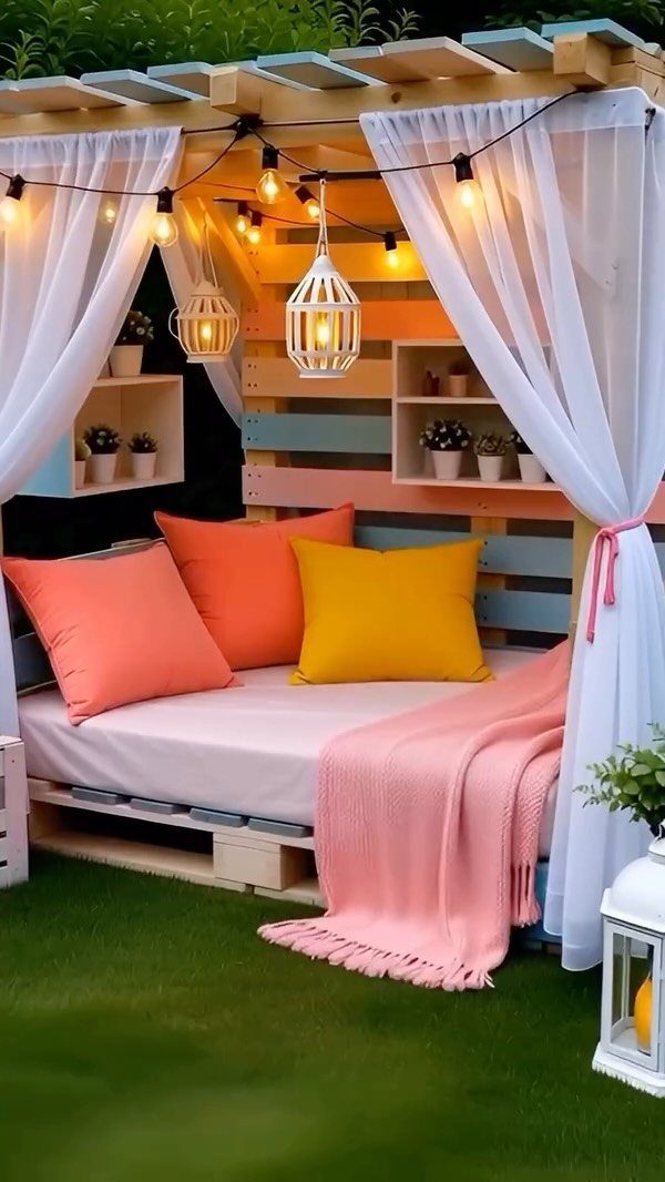 Pallet daybed with colorful pillows and white curtains under a lit canopy.