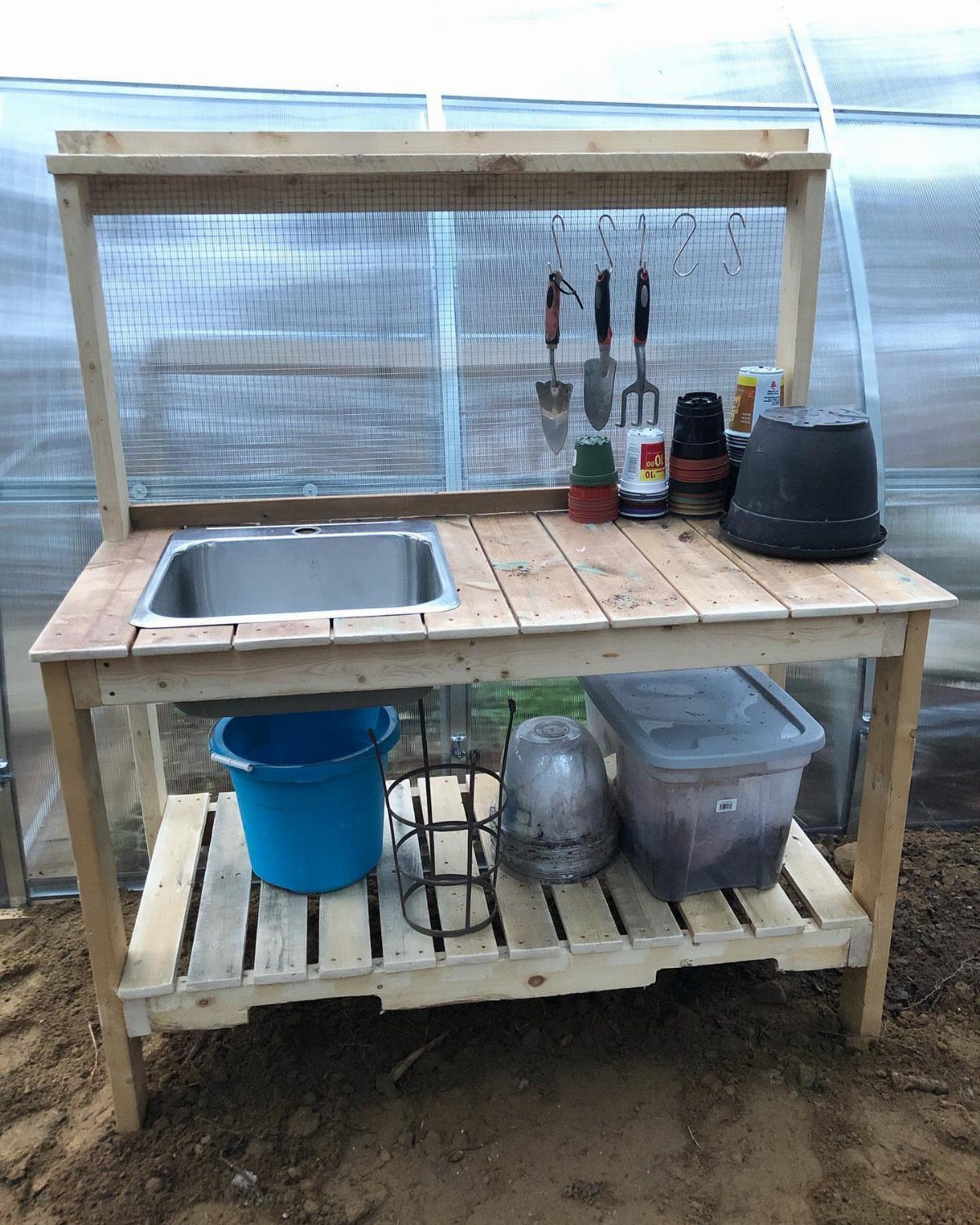 Pallet wood potting bench with metal sink and gardening tools in a greenhouse.