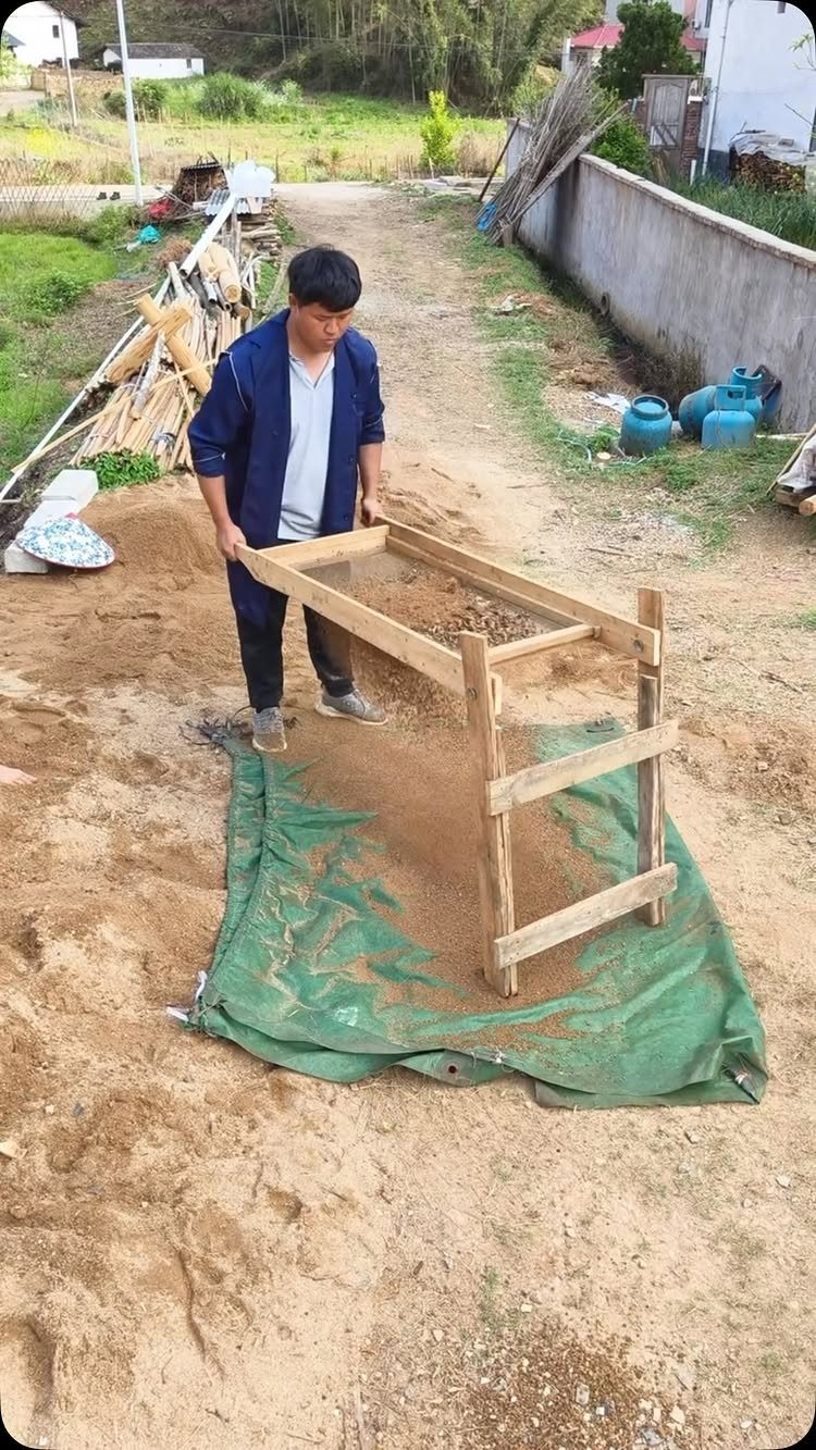 Man using a wooden soil sifter to filter dirt onto a green tarp.
