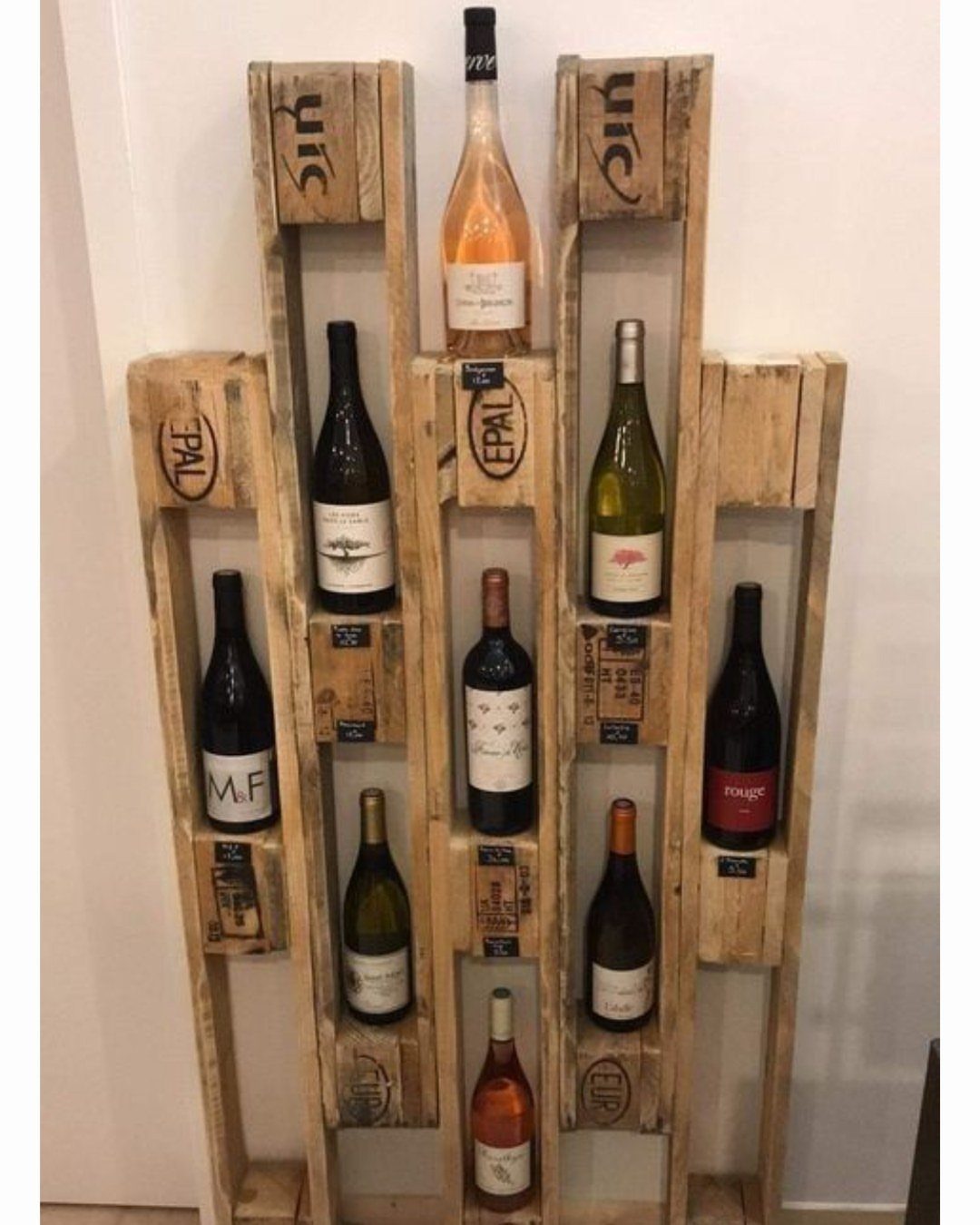 Rustic pallet wood wine rack holding several bottles of wine against a white wall.