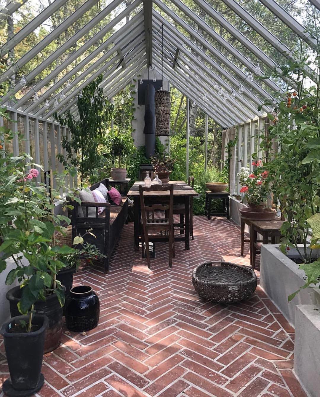 Greenhouse interior with red brick herringbone flooring, wooden furniture, and lush potted plants.