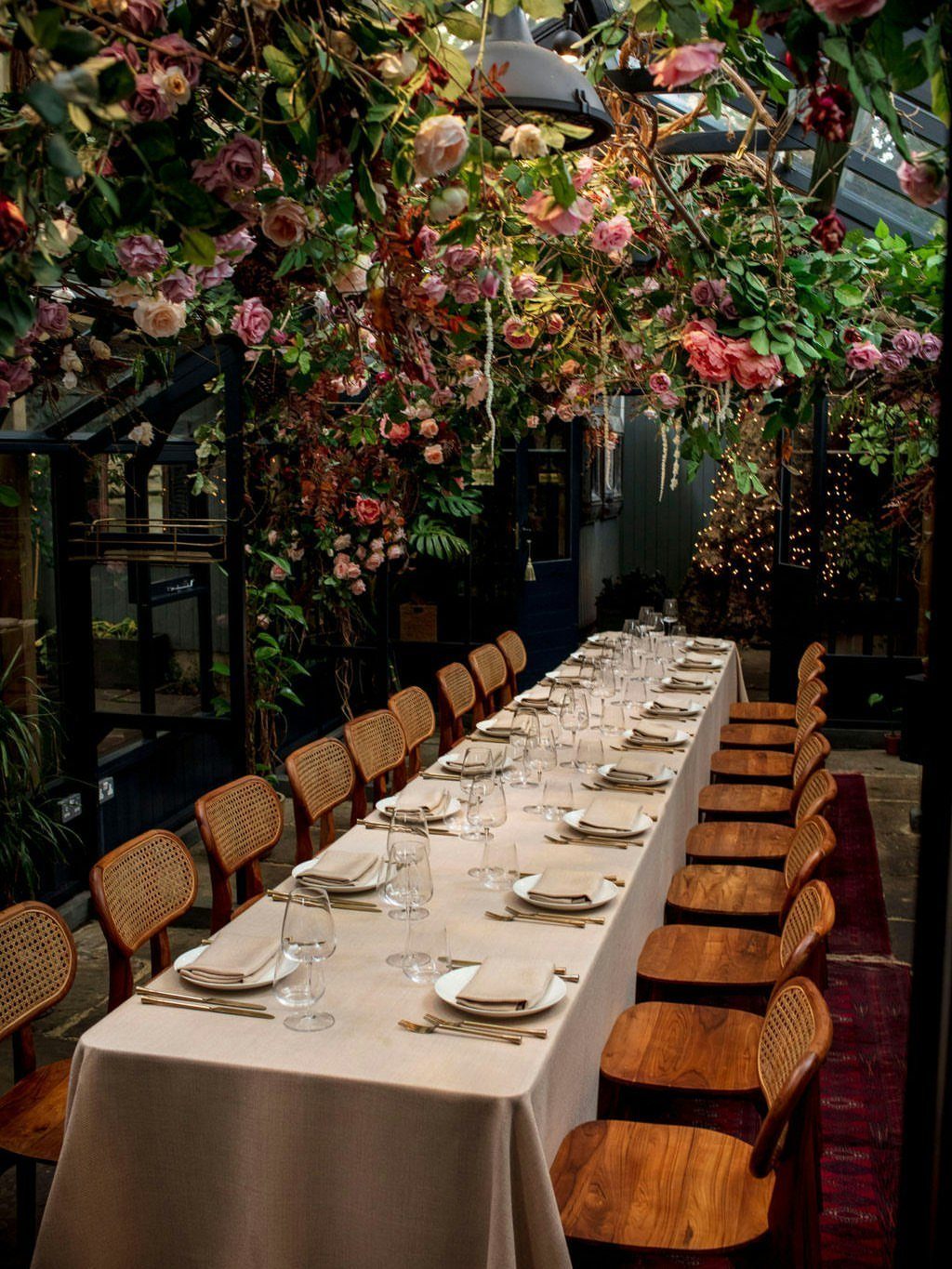 Long dining table set for a meal under a canopy of pink roses.