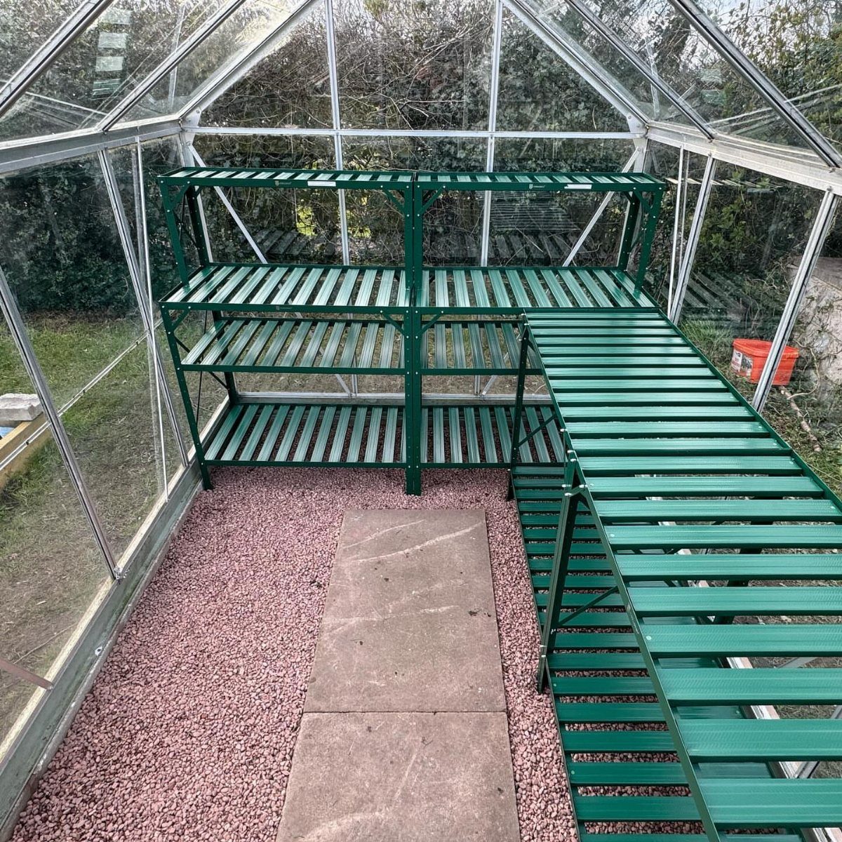 Green slatted shelving and table inside a glass greenhouse with gravel flooring.