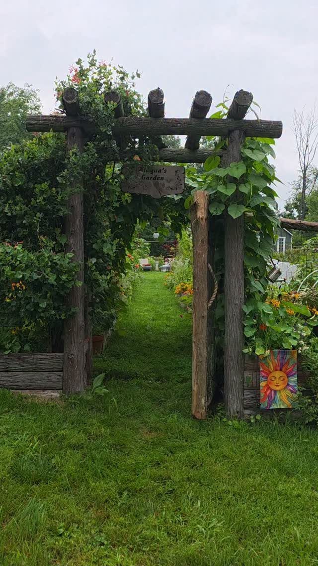 Wooden pergola archway with green vines leading into a lush vegetable garden path.