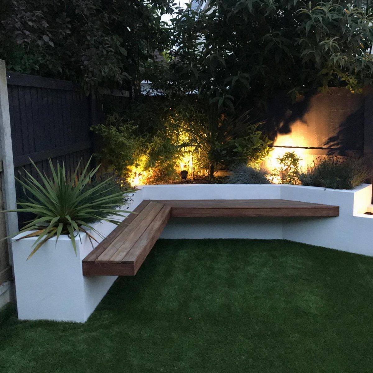 Raised Beds Garden: 25 Ideas from Wood, Metal, and Stone 15 White L-shaped raised garden bed with a wooden bench seat in a lit backyard.