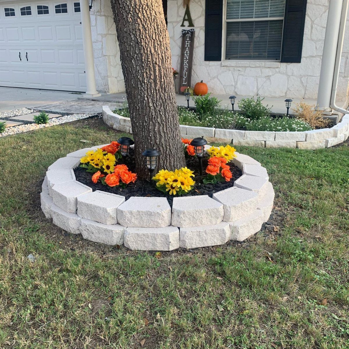 Circular stone raised flower bed with orange and yellow flowers around a tree trunk.