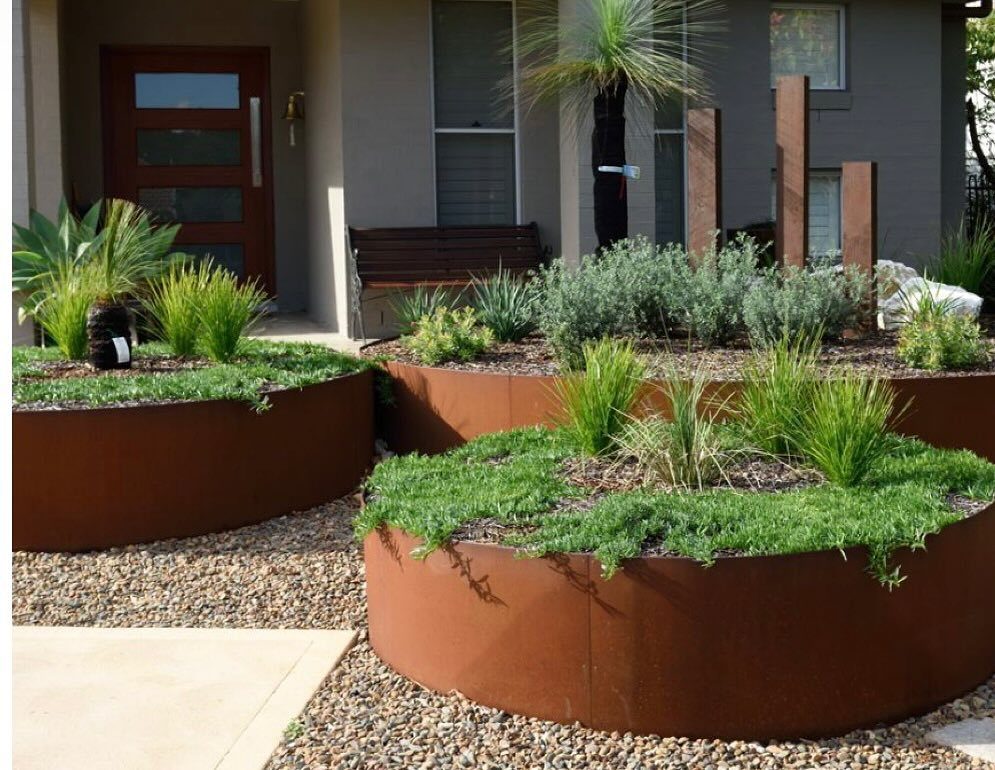 Circular rusted steel raised garden beds with green plants in a gravel yard.