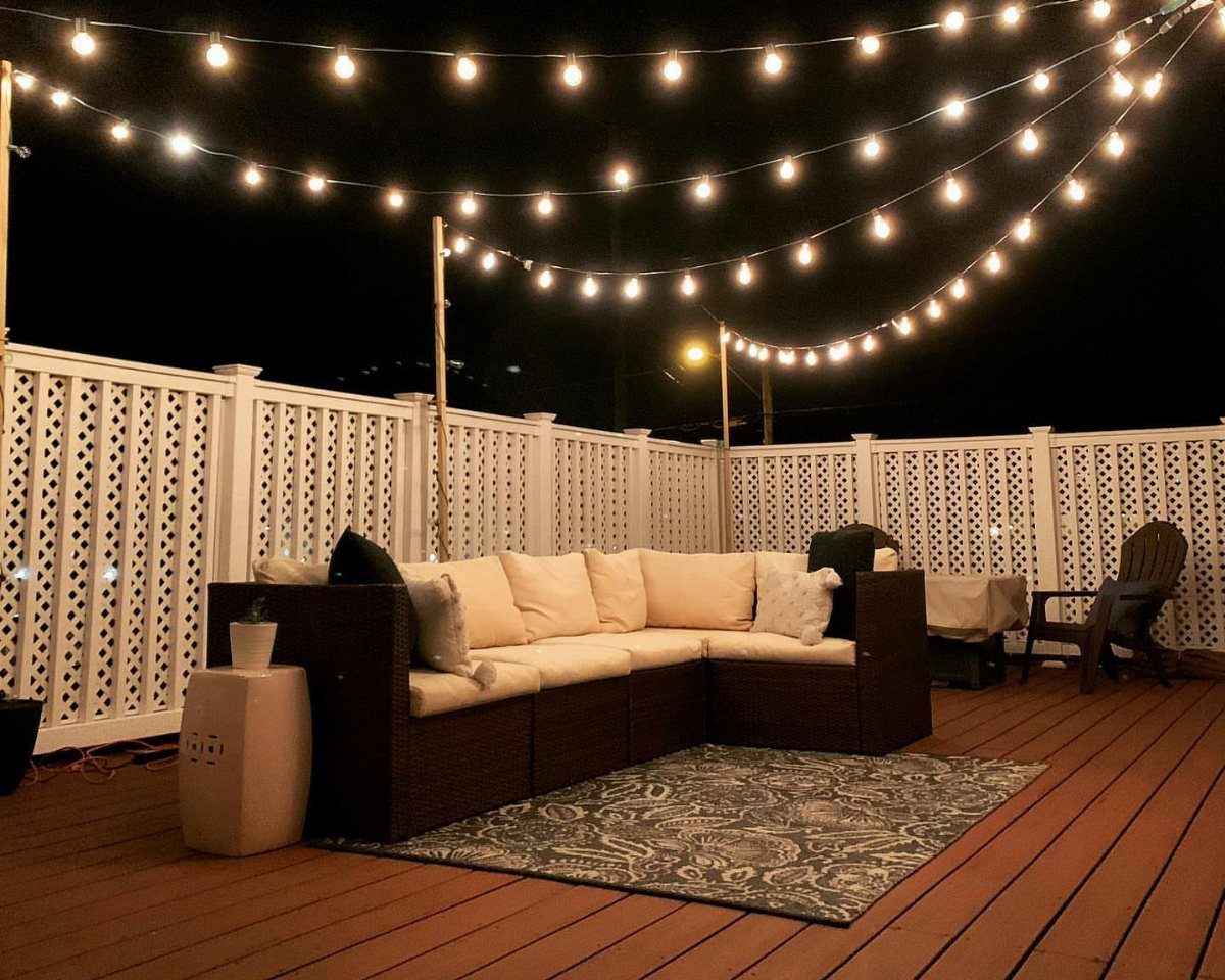 Rooftop deck at night with a wicker sectional sofa under glowing string lights.