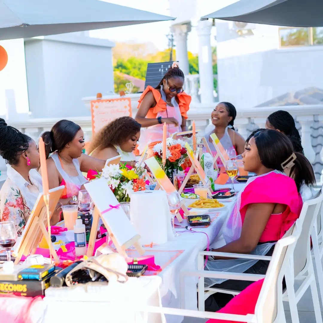 Women painting on canvases at a white table on a sunny rooftop terrace.