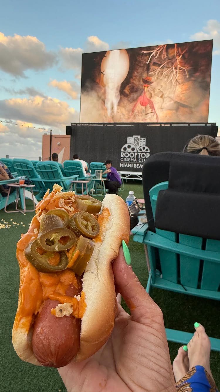 Hot dog with jalapeños held in front of a rooftop cinema screen and lounge chairs.