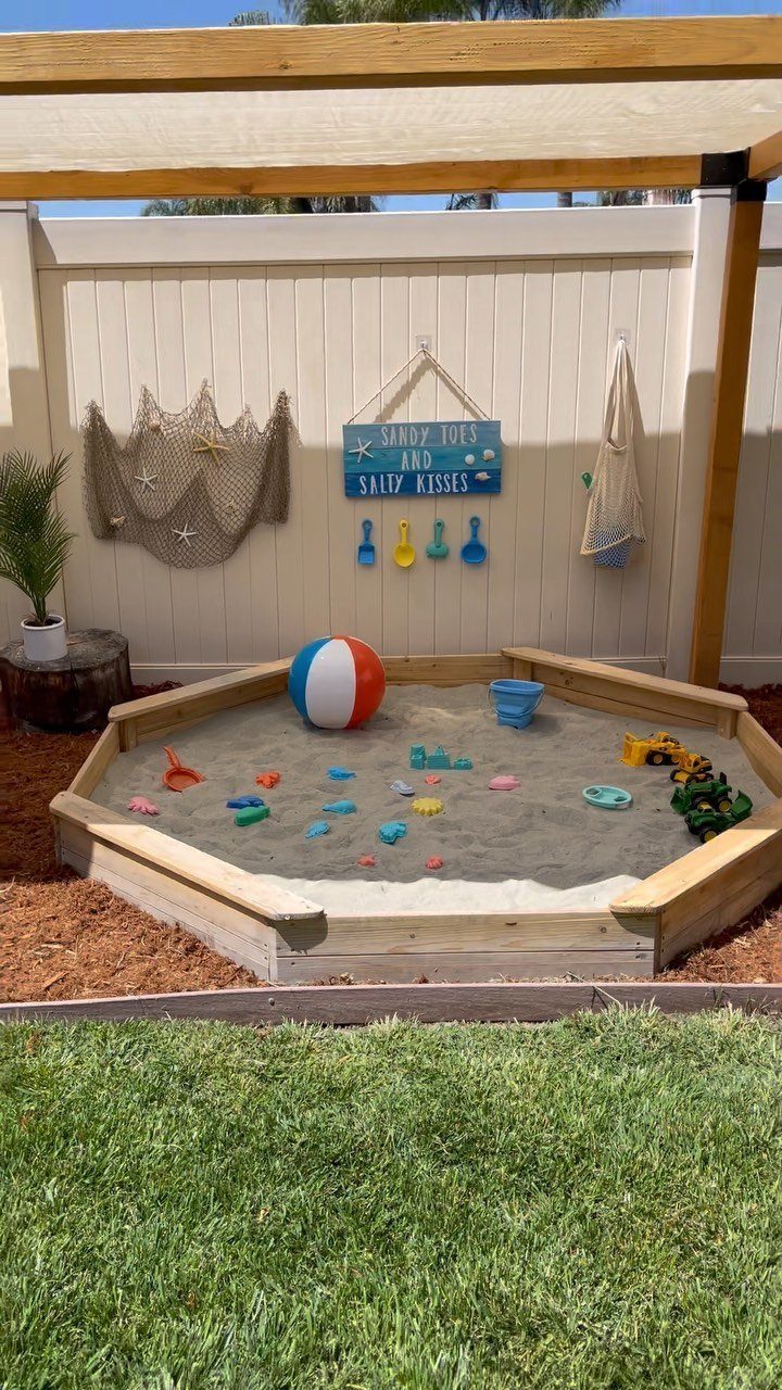 Wooden octagonal sandbox with toys and a beach ball under a wooden pergola.