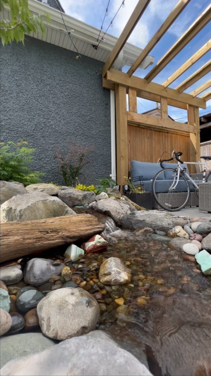 Small natural garden pond with river stones and a log under a wooden pergola.