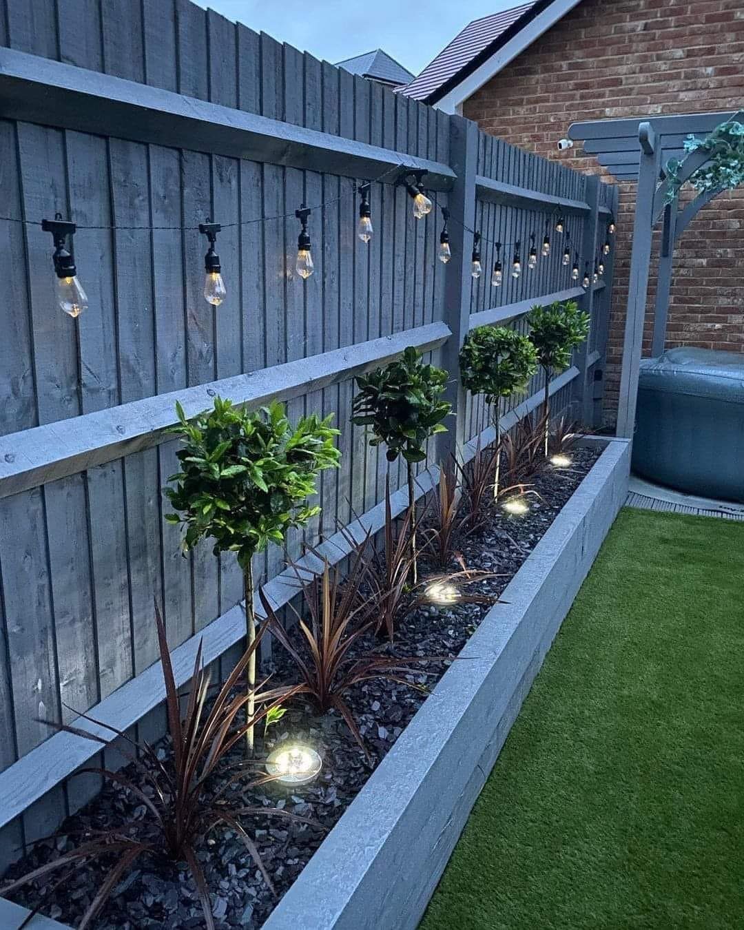 String lights draped across a grey wooden fence in a narrow landscaped garden border.