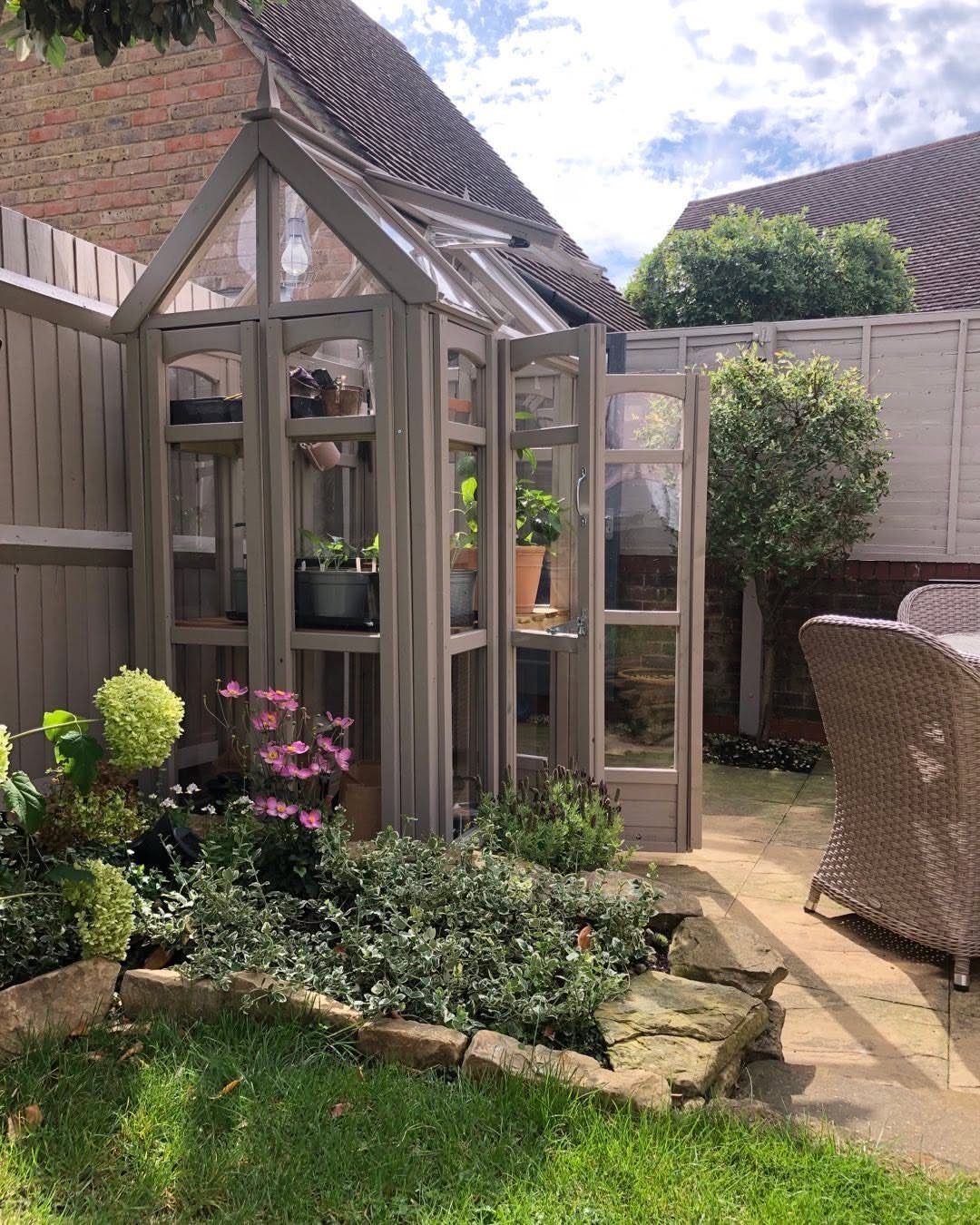 Grey glass greenhouse with potted plants in a garden next to a brick house.