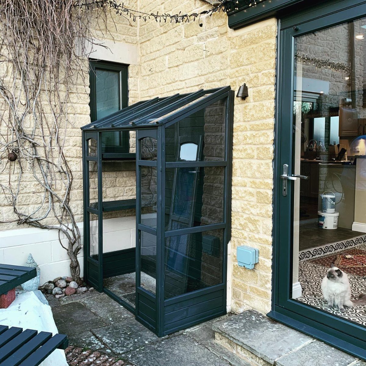 Small dark grey glass greenhouse leaning against a light stone house wall.