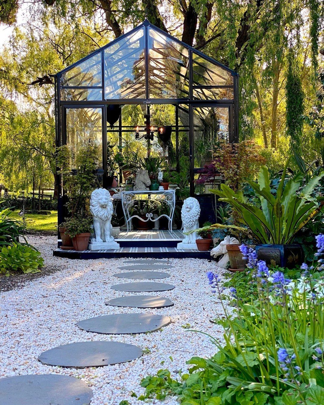 Glass greenhouse with black frame and white gravel path in a lush garden