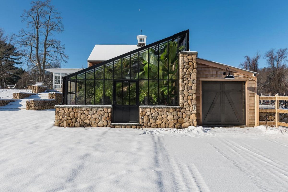 Modern glass greenhouse with stone base attached to a wooden building in snow.