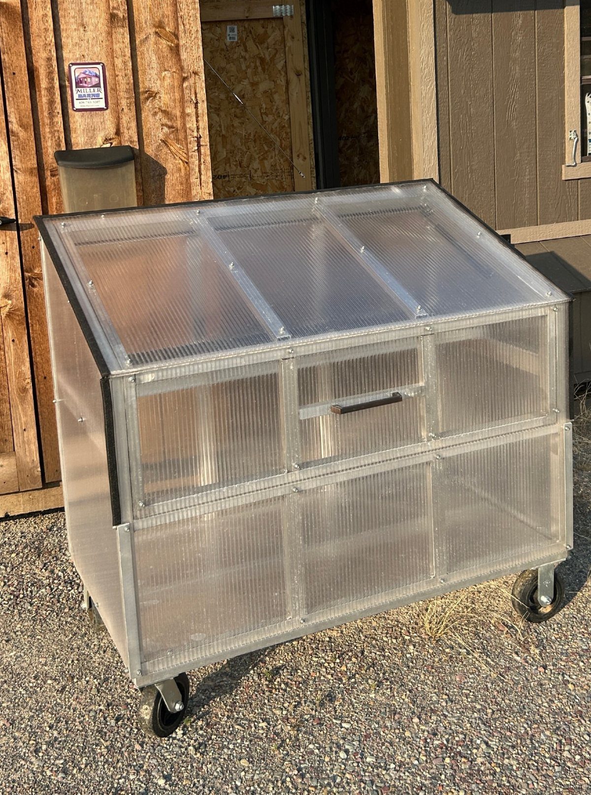 Small polycarbonate greenhouse on wheels sitting on a gravel ground next to wood walls.