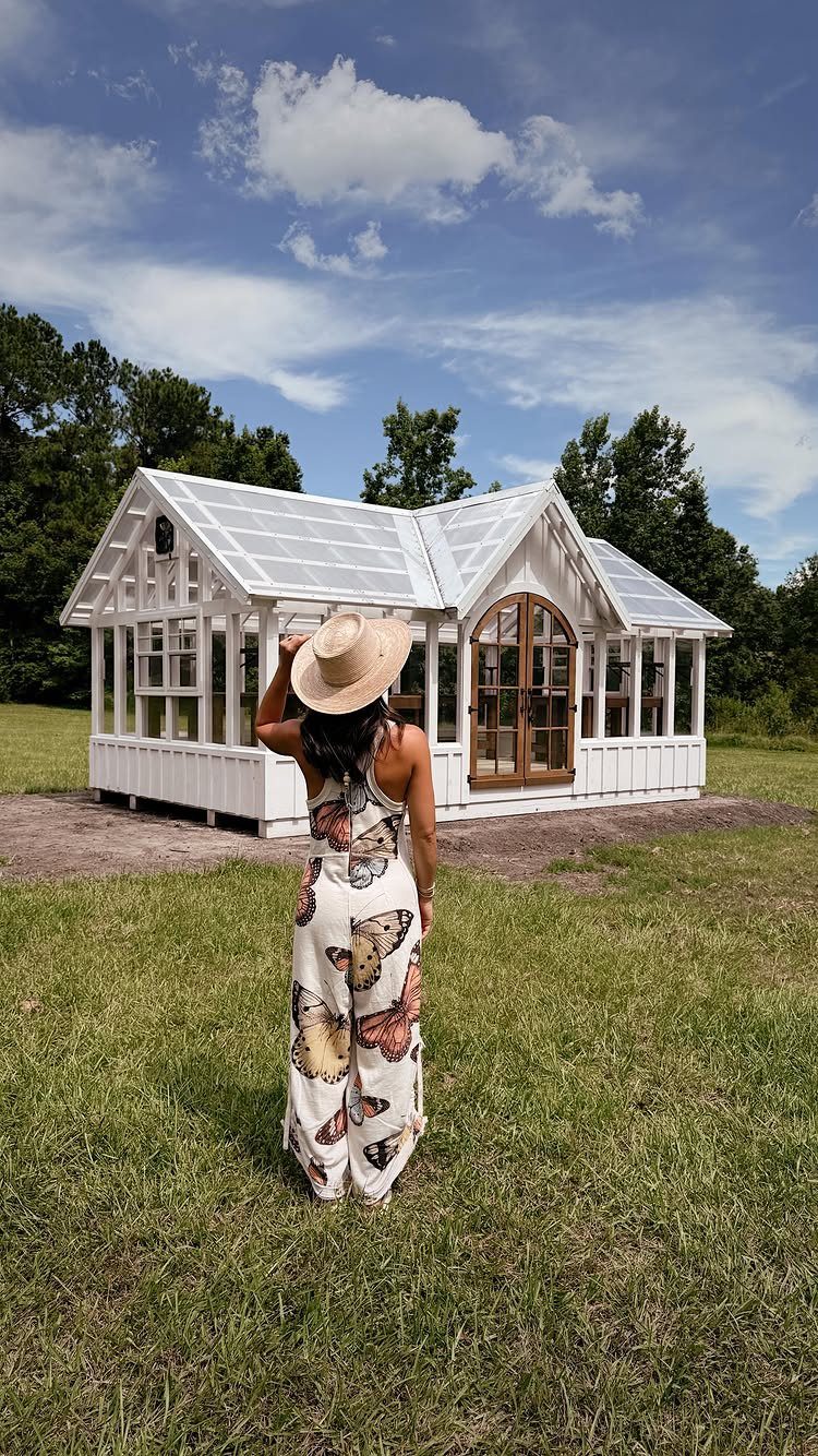 Woman in butterfly jumpsuit standing before a white modern gabled greenhouse in a field.