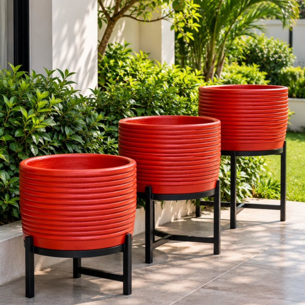 Three red ribbed planters on black stands arranged on a sunny stone patio.