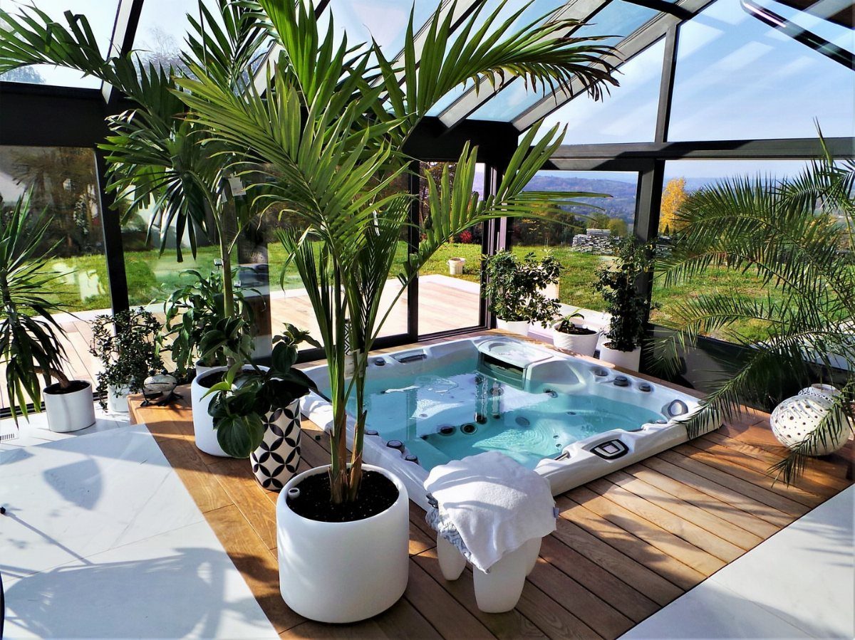 Hot tub on a wooden deck inside a glass sunroom with potted palm trees.