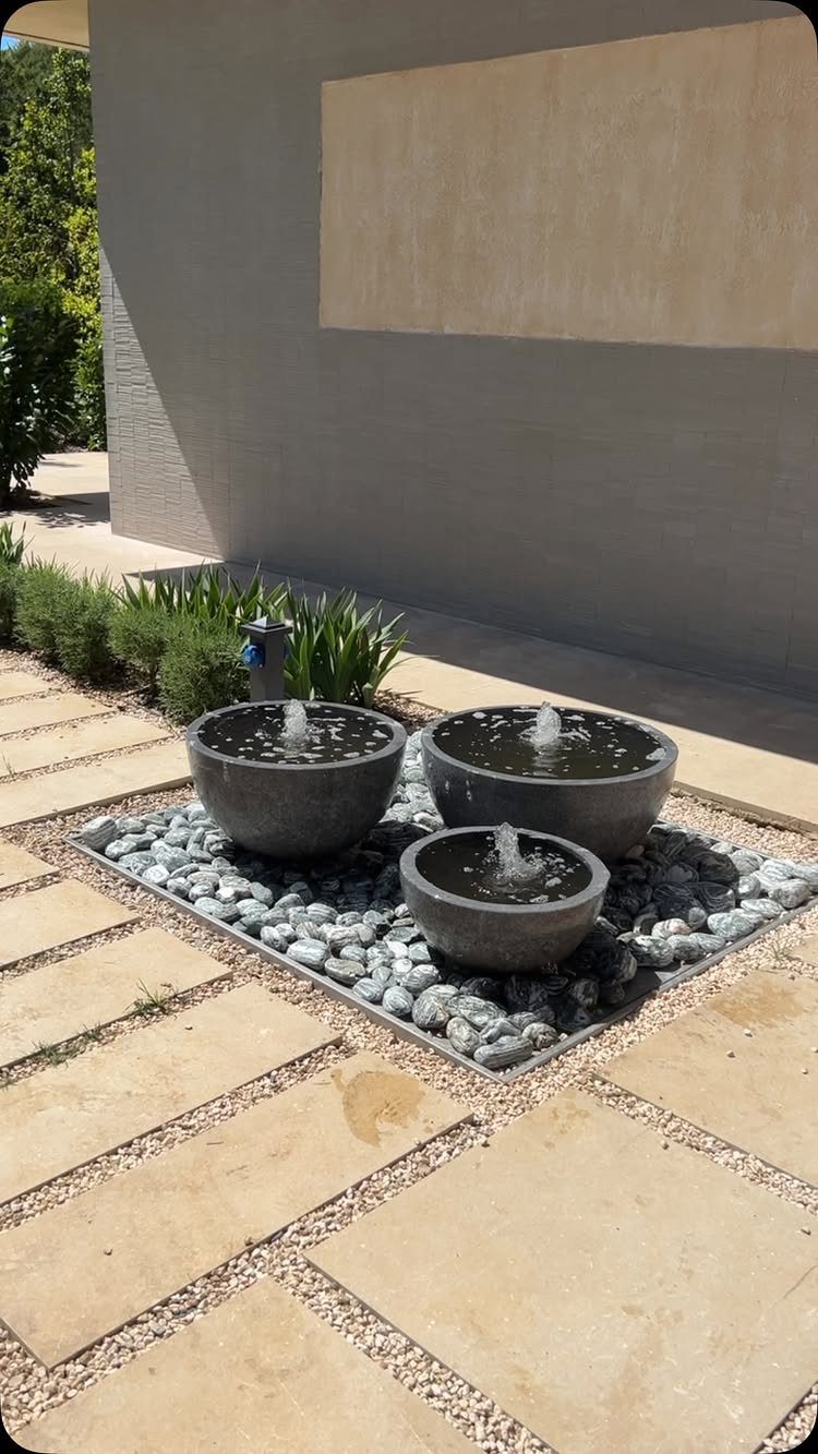 Three stone water bowls with fountains set in grey pebbles on a stone patio.